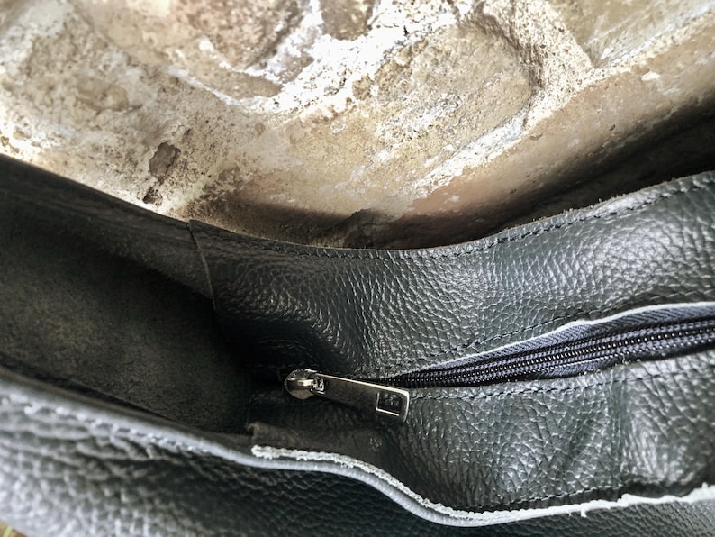 May include: Close-up of a dark gray leather bag with a textured surface. The bag has a silver zipper and a contrasting white lining. The image shows the inside of the bag, with a glimpse of the opening.