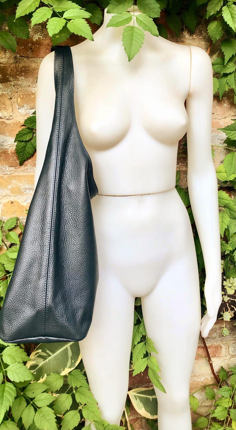 May include: A dark teal leather shoulder bag with a textured surface. The bag is displayed on a white mannequin against a backdrop of brick and greenery. The bag's strap is visible.
