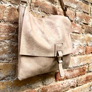 May include: A light beige leather crossbody bag with a flap closure and a buckle. The bag has a worn, vintage look and is hanging on a brick wall.
