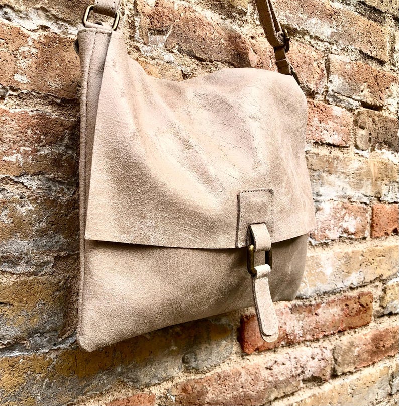 May include: A light beige leather crossbody bag with a flap closure and a buckle. The bag has a worn, vintage look and is hanging on a brick wall.