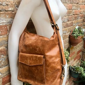 Op de afbeelding: Een bruine leren rugzak met een klep sluiting en een voorvak. De tas heeft een verweerde look en wordt gedragen door een mannequin.