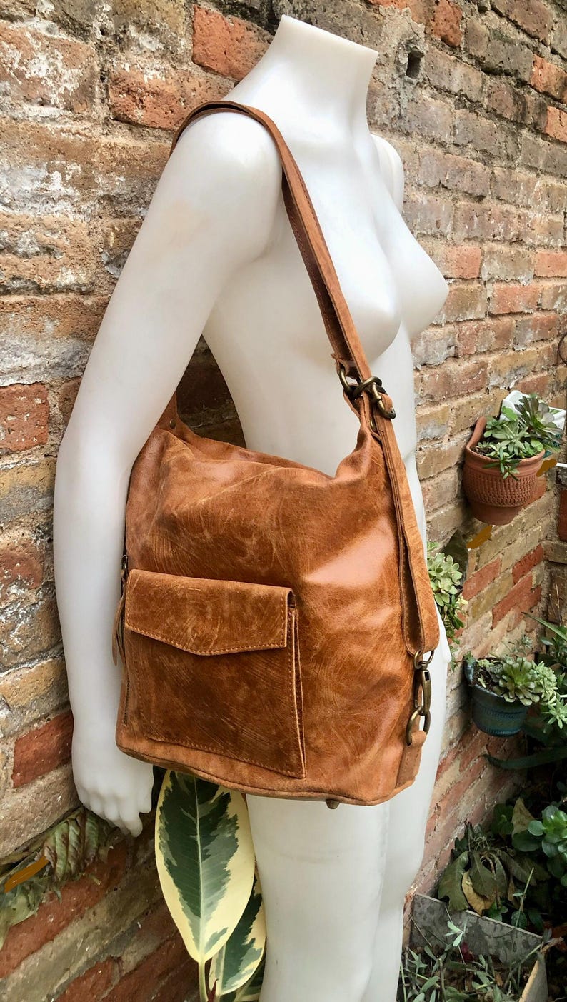 Op de afbeelding: Een bruine leren rugzak met een klep sluiting en een voorvak. De tas heeft een verweerde look en wordt gedragen door een mannequin.