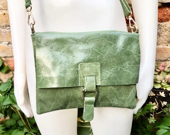 May include: A green leather crossbody bag with a flap closure and a buckle. The bag has a long, adjustable strap and gold-colored hardware. The bag is displayed on a white mannequin.