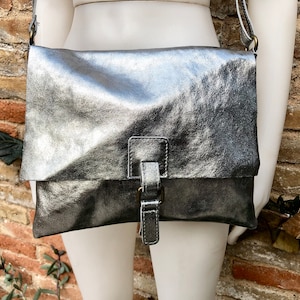 May include: Silver crossbody bag with a flap closure and adjustable strap. The bag features a buckle detail on the front and a metallic finish. The bag is displayed on a mannequin.