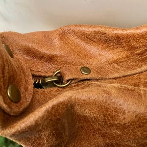 Op de afbeelding: Close-up van een bruine leren tas met een rits en metalen hardware. De tas heeft een versleten, verweerde look.
