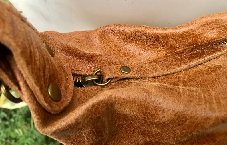 Op de afbeelding: Close-up van een bruine leren tas met een rits en metalen hardware. De tas heeft een versleten, verweerde look.