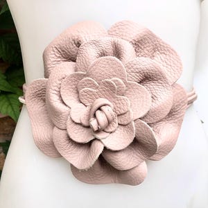 May include: A blush pink leather belt featuring a large, dimensional flower design. The flower is the focal point, with multiple overlapping petals creating a textured look. The belt is displayed on a mannequin.