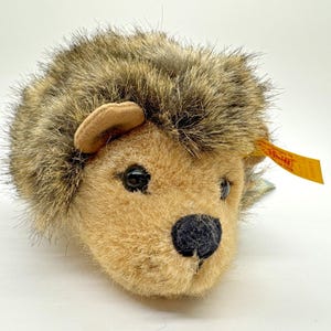 Puede incluir: Un juguete de peluche de erizo con cuerpo beige, púas marrones y beige, y una nariz negra. El juguete tiene ojos pequeños y negros y una oreja beige. Una etiqueta amarilla está adjunta.