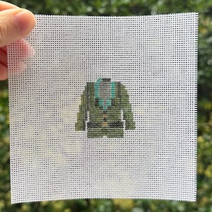 May include: A small, square piece of white canvas with a detailed cross-stitch design of a green jacket with a teal collar. The jacket has gold-colored buttons and a pocket. The background is a blurred view of green foliage.