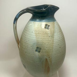 May include: A handmade ceramic pitcher with a dark blue rim and handle, transitioning to a light blue and beige body. The pitcher features a unique spout design and decorative markings. The overall shape is rounded with a sturdy handle.