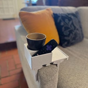 Puede incluir: Una bandeja gris claro para el brazo del sofá con un portavasos incorporado y almacenamiento para el control remoto. Un control remoto negro y una taza marrón oscuro están en la bandeja. La bandeja está sujeta al brazo de un sofá gris. Un cojín naranja y un cojín estampado están en el sofá.
