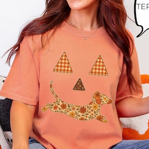 May include: A terracotta-colored t-shirt featuring a graphic of a dachshund dog with a floral pattern, positioned to form a smile, with two plaid triangles for eyes and one for a nose. The shirt includes a speech bubble graphic with the text TERRACOTTA.