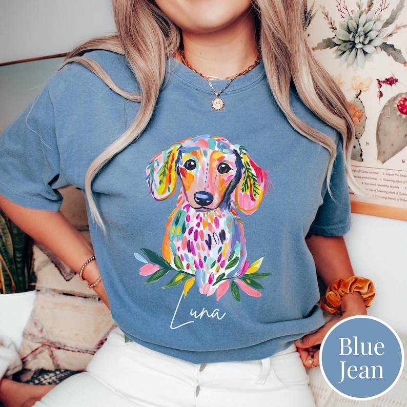 May include: A blue jean colored t-shirt featuring a colorful watercolor illustration of a dachshund dog. The dog has a vibrant coat with pink, yellow, and blue accents. The name "Luna" is written in cursive below the dog.