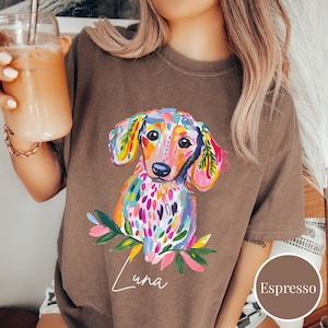 May include: Espresso-colored t-shirt featuring a colorful, artistic dachshund illustration. The dog is depicted with vibrant hues of pink, blue, yellow, and green. The name "Luna" is written in cursive below the dog. The shirt is a comfortable, casual style.