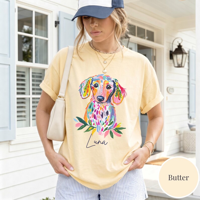May include: Butter-colored t-shirt featuring a colorful watercolor dachshund illustration with the name "Luna" below. The dog has a vibrant mix of colors, including pink, blue, and green. The shirt is a casual style.