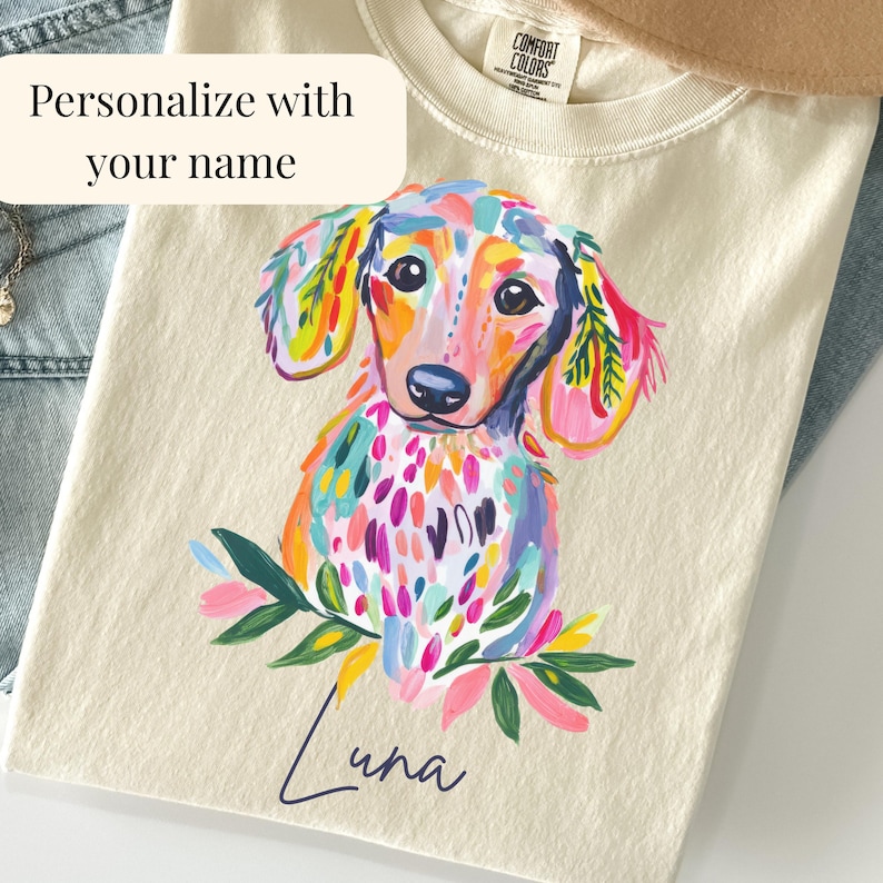 May include: Cream-colored t-shirt featuring a colorful dachshund illustration. The dog is painted with vibrant hues of pink, blue, yellow, and green. The name "Luna" is written in cursive below the dog. Text on the shirt reads "Personalize with your name."