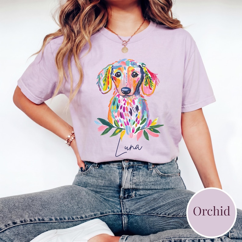 May include: A light purple t-shirt featuring a colorful watercolor dachshund illustration. The dog has a rainbow-colored coat and is surrounded by floral accents. The name "Luna" is written in cursive below the dog.