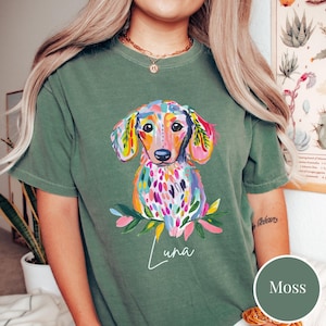 May include: Moss green t-shirt featuring a colorful watercolor dachshund illustration. The dog has a vibrant coat of pink, blue, yellow, and green. The name "Luna" is written in cursive below the dog. The shirt is a comfortable, casual style.