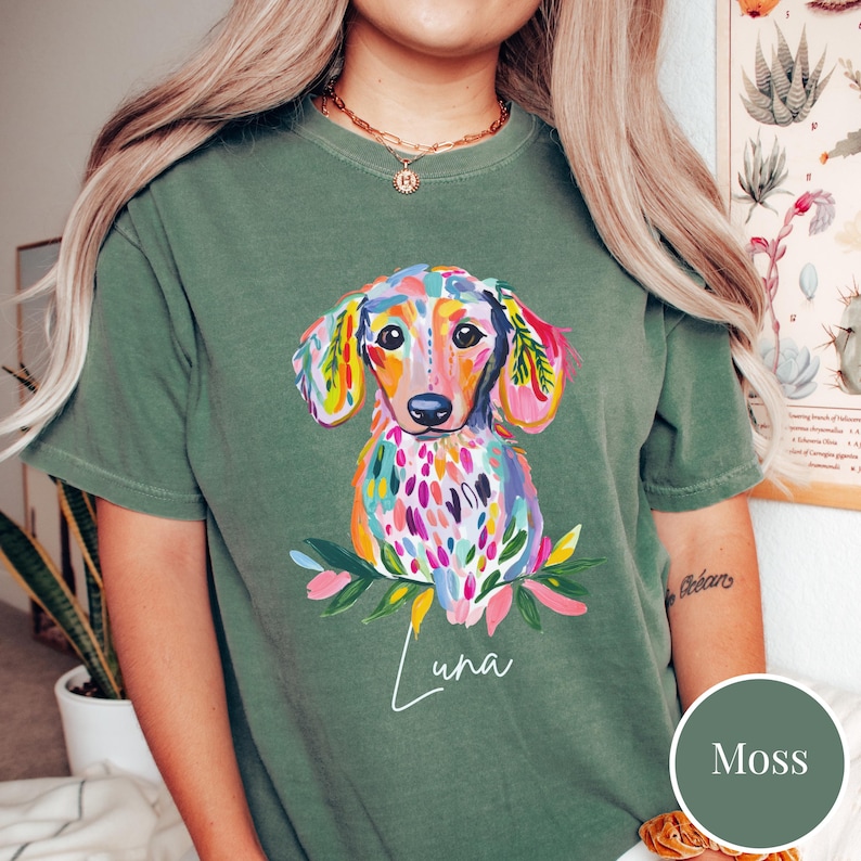 May include: Moss green t-shirt featuring a colorful watercolor dachshund illustration. The dog has a vibrant coat of pink, blue, yellow, and green. The name "Luna" is written in cursive below the dog. The shirt is a comfortable, casual style.