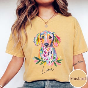May include: Mustard-colored t-shirt featuring a colorful dachshund dog illustration. The dog has a vibrant design with pink, blue, green, and yellow accents. The name "Luna" is written in cursive below the dog. The shirt is a relaxed fit.