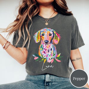 May include: A dark gray t-shirt featuring a colorful watercolor dachshund illustration. The dog is depicted with vibrant hues of pink, yellow, blue, and green. The name "Luna" is written in cursive below the dog. The shirt is a Pepper color.