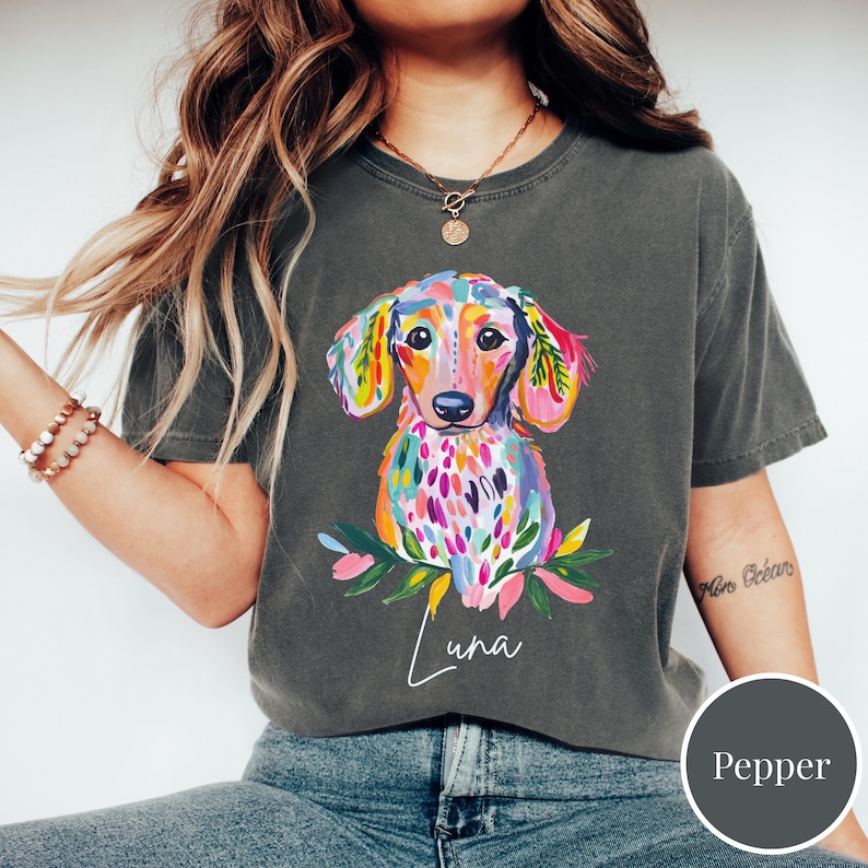 May include: A dark gray t-shirt featuring a colorful watercolor dachshund illustration. The dog is depicted with vibrant hues of pink, yellow, blue, and green. The name "Luna" is written in cursive below the dog. The shirt is a Pepper color.
