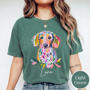 May include: A light green t-shirt featuring a colorful watercolor dachshund illustration. The dog has a vibrant mix of colors, including pink, yellow, and blue. The name "Luna" is written in cursive below the dog.