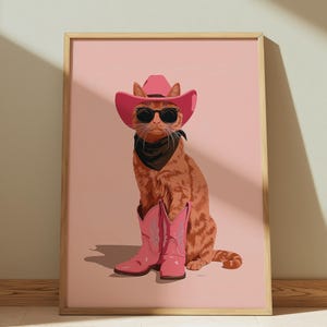 May include: A framed print featuring an orange tabby cat wearing a pink cowboy hat, sunglasses, a black bandana, and pink cowboy boots. The cat is sitting against a pink background. The artwork has a whimsical and playful theme.