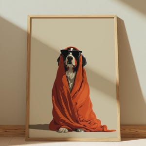 Op de afbeelding: Een ingelijste print met een hond met een zonnebril en gewikkeld in een oranje deken. De hond heeft zwart-witte vacht en zit tegen een neutrale achtergrond. Het kunstwerk zit in een houten lijst.