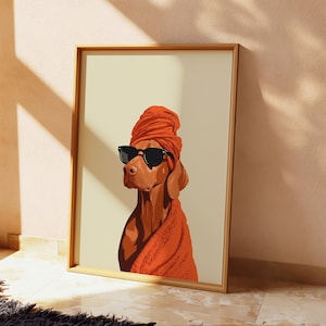 May include: A framed art print depicting a brown dog wearing black sunglasses and an orange head wrap and shawl. The dog is set against a cream-coloured background. The artwork is in a wooden frame.