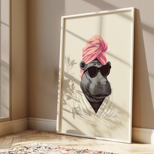 May include: A framed art print featuring a stylized hippo wearing black sunglasses and a pink turban. The hippo is depicted in a white robe against a cream-colored background. The artwork is displayed in a white frame.