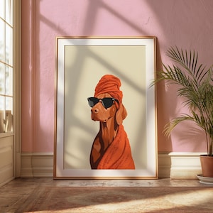 May include: A framed art print depicting a dog in black sunglasses, an orange head wrap, and a matching towel. The dog is a reddish-brown colour. The artwork is presented in a gold frame against a cream background, creating a stylish aesthetic.