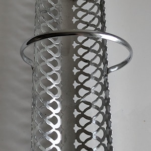 Puede incluir: Un portautensilios de metal plateado con un diseño calado decorativo. El soporte tiene forma cilíndrica con un asa y un borde superior festoneado. El patrón calado es una serie repetida de formas entrelazadas.