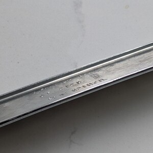 May include: A chrome-finished, rectangular automotive trim piece with two mounting studs. The trim has a slightly curved design and a polished surface. The image is a close-up shot of the trim piece.