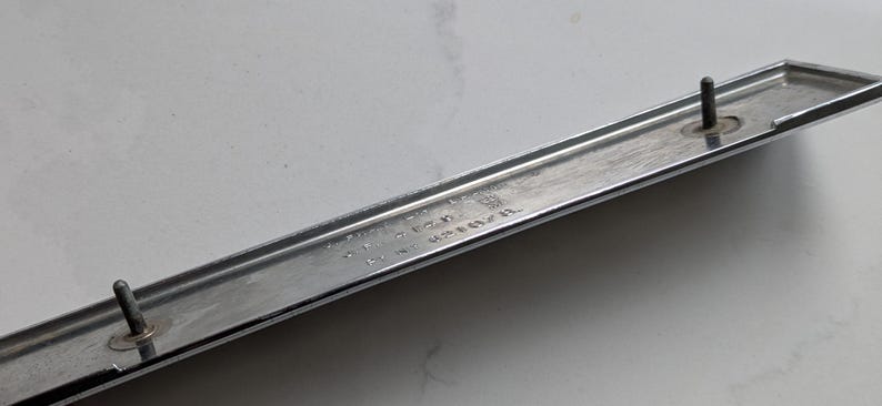 May include: A chrome-finished, rectangular automotive trim piece with two mounting studs. The trim has a slightly curved design and a polished surface. The image is a close-up shot of the trim piece.