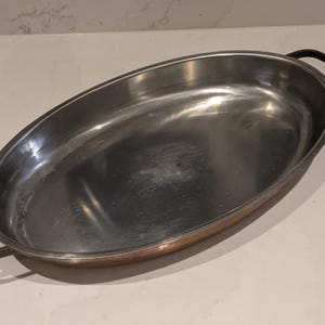 Può includere: Piatto ovale in metallo con bordi e manici color bronzo. L'interno ha una finitura in metallo spazzolato. I manici sono di un colore bronzo più scuro. Il piatto è vuoto e poggia su un piano di lavoro bianco.