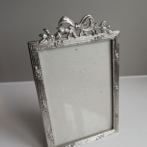 May include: Silver-colored rectangular picture frame with a bow and floral design at the top. The frame has a vertical ribbed pattern and decorative accents on the sides. The interior is a light gray color.