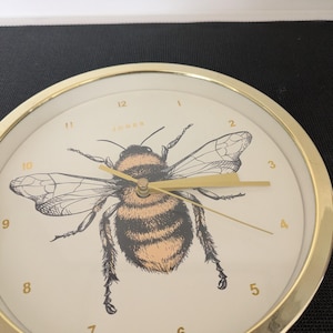 May include: A round wall clock with a gold-colored metal frame. The clock face is cream-colored with a large illustration of a bee. The bee is black, orange, and tan. Gold-colored hands and numbers complete the design. The word "JONES" is printed on the clock face.