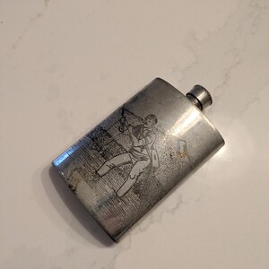 May include: A rectangular, silver-toned flask with a screw-on cap. The flask features an etched illustration of a person in period clothing. The flask has a slightly worn appearance, suggesting age and use. The flask is a vintage item.