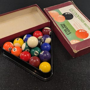 May include: A vintage set of Guinness Brothers snooker and pool balls. The balls are arranged in a black triangle rack, with a white cue ball and numbered balls in various colours. The box is maroon with the Guinness Brothers logo.