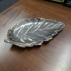 Puede incluir: Un plato decorativo plateado con forma de hoja. El plato tiene vetas detalladas y una superficie texturizada, que se asemeja a una hoja real. El plato está colocado sobre una superficie de madera marrón.