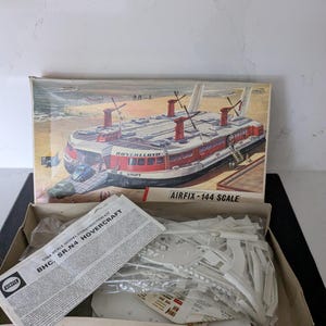 May include: An open box containing an Airfix 1/144 scale model construction kit of a BHC SR.N4 Hovercraft. The box art depicts the Hovercraft Swift on a beach. The box is open, revealing unassembled white plastic model parts and instructions.