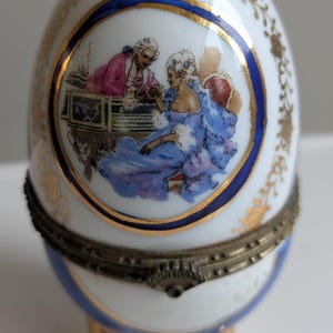 May include: A decorative egg-shaped trinket box. The white porcelain egg features a blue and gold trim, with a painted scene of figures in period clothing. The egg rests on three gold-colored feet and has a hinged metal base.