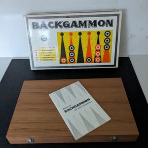Op de afbeelding: Een vintage Backgammon-spelset. De doos toont de naam van het spel in vette letters, met een kleurrijk ontwerp van driehoeken en cirkels. Het houten spelbord is open, met een boekje getiteld "Regels van Backgammon" door John Davidson.