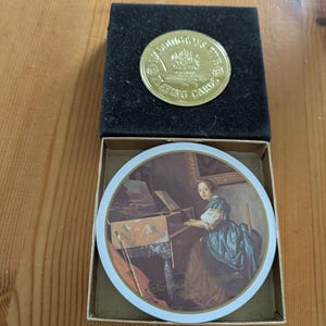 May include: A gold-colored coin with the text "SOMETHING'S PLAYING CARDS" in a black velvet box. Below, a round card features a painting of a woman playing a harpsichord. The card is in a cardboard box.