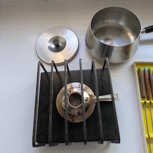 May include: A vintage fondue set with a stainless steel pot, lid, and burner. The burner sits on a black metal stand. A box of fondue forks is also visible.