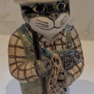 Può includere: Statuina in ceramica di un gatto vestito da pescatore. Il gatto indossa una camicia a quadri, una salopette verde e un cappello da pescatore. Tiene una rete da pesca e una canna da pesca. La statuina ha un design dettagliato dipinto a mano.