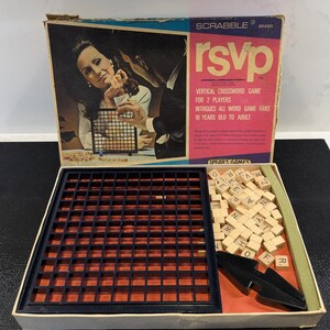 Può includere: Gioco da tavolo Scrabble RSVP vintage. La scatola presenta un design retrò con il titolo del gioco in rosa e bianco. All'interno, sono visibili una griglia di gioco blu scuro, tessere di lettere in legno e un pezzo di gioco nero.
