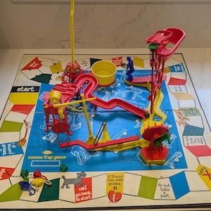 May include: A vintage Mouse Trap board game with a blue game board and colorful plastic components. The game includes a yellow bucket, red slides, and a yellow and red seesaw. The board game has the word "start" printed on it.