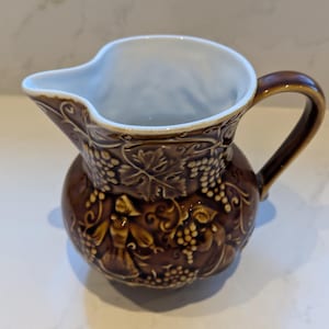 Può includere: Una brocca in ceramica marrone con interno bianco e manico curvo. La brocca è decorata con un motivo a rilievo di viti e grappoli d'uva. Il beccuccio è progettato per versare facilmente.